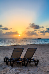 Two beach chairs are facing the ocean, with the sun setting in the background