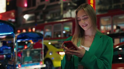Portrait of blonde woman in casual outfit looking at mobile phone screen in night city. Positive female reads messages in online chats via smartphone against urban traffic in evening - Powered by Adobe