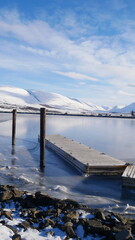 Pomost w zatoce. Zima w Akureyri. Zamarznięta zatoka z drewnianym pomostem. Fiord, góry, zima na Islandii.  © onlytraveler