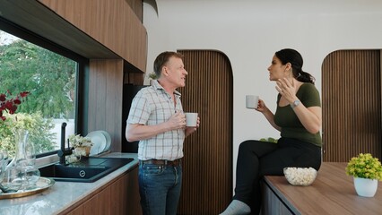 Lovely old marriage couple happily talking about good event or news in kitchen while holding coffee cup to drink. Cheerful grandfather and grandmother have happy relationship and family. Divergence.