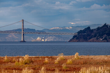 The photograph captures the serene morning atmosphere with calm waters of the San Francisco Bay, the iconic bridge structure, and the historic Alcatraz Island.