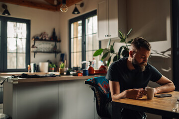Bearded man using smartphone and drinking coffee in kitchen