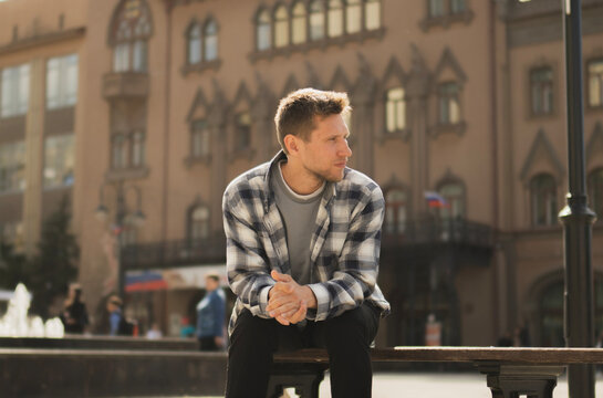 A man in a checked shirt sits thoughtfully on a bench near a striking cathedral structure as the sun sets, casting warm light