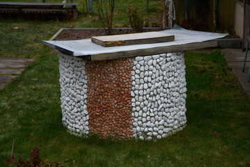 A decorative outdoor structure made of white and red pebbles with a flat metal top, set on a grassy backyard.