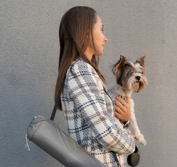 A woman stands outdoors holding a small dog close to her chest while casually wearing a trendy...