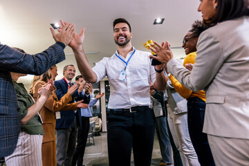 Young businessman receiving prize cup, proud colleagues applauding for him and cheering.