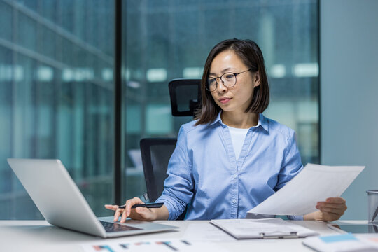 Asian woman businesswoman engaged in work at her desk, using a laptop and reviewing documents. The office setting highlights productivity and professionalism, reflecting a focused and organized work