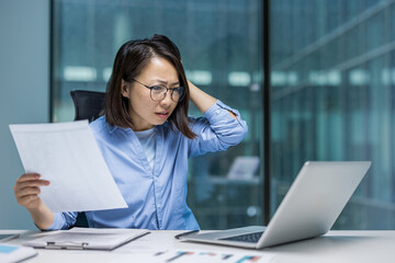 An Asian businesswoman appears frustrated looking at her laptop and holding documents. The scene...