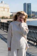 Portrait of beautiful smiling long haired blond caucasian woman in white clothes walking on city street embankment in a sunny autumn day. Soft focus. Fashion and happy people theme.