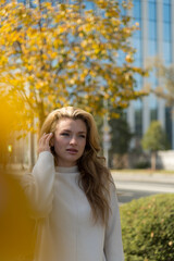 Portrait of beautiful smiling serene long haired blond caucasian woman in white sweater standing outdoors on city street in a sunny autumn day by tree with yellow leaves. Soft focus. People theme.