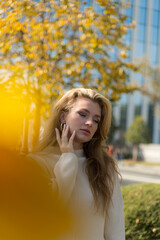 Fototapeta premium Portrait of beautiful smiling serene long haired blond caucasian woman in white sweater standing outdoors on city street in a sunny autumn day by tree with yellow leaves. Soft focus. People theme.