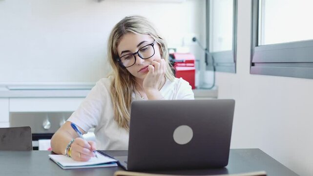 Millennial blonde girl sit at desk in living room study on laptop making notes, concentrated young woman work on computer write in notebook, take online course or training at home, education concept
