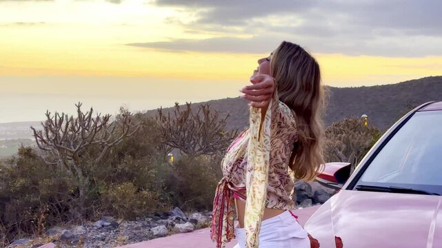 Young beautiful woman enjoying summer and having fun sitting on the hood of the car opening arms feeling good and free. Freedom concept and happy lifestyle at sunset
