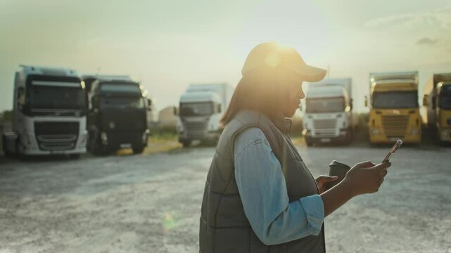 Side camera view of African American woman walking in front of row of parked trucks. Typing on screen of smartphone with one hand while holding coffee cup with other. Bright sunshine in background.