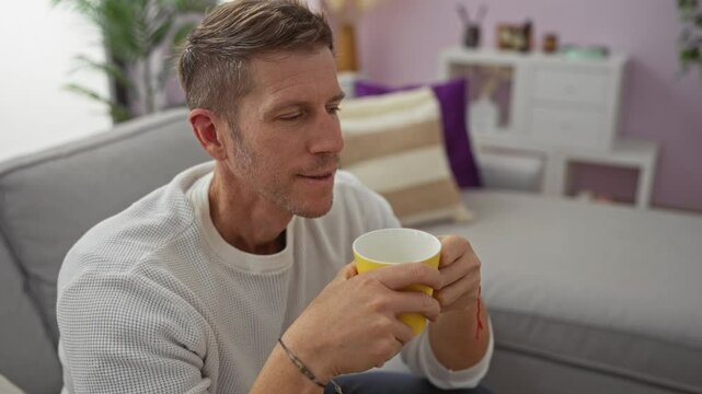 Young man enjoying coffee in cozy living room with modern decor, relaxing on sofa, holding yellow cup, and wearing casual white sweater.