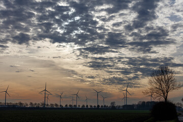 Silhouetted wind turbines stand against a dramatic, cloudy sunset sky.  A lone tree stands in the foreground.