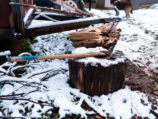 The axe lies on a wooden stump with a pile of chopped firewood on the snow-covered ground. Heating the house with wood.