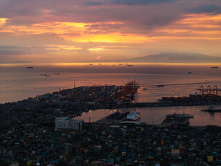 Manila's lively port area at sunset, featuring cargo ships dotted across the horizon against a dramatic sky with colorful clouds, and city lights twinkling below