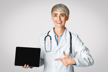 Online making appointment and consultation. Smiling woman doctor points finger at tablet with blank screen, isolated on light background, studio shot