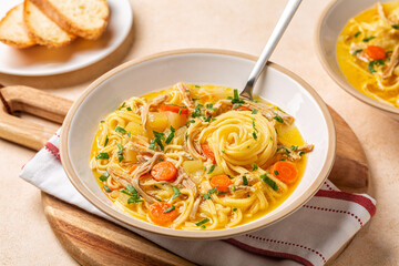 Dinner with chicken noodle soup in a white plate, on a wooden board. Beige table surface.