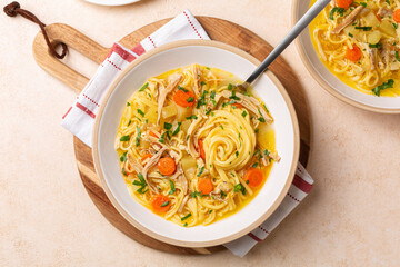 Chicken noodle soup in a white plate, on a wooden board. Directly above on a beige table.