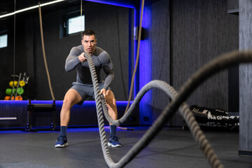 An athletic man highlights strength and power with continuous battle rope movements in a state-of-the-art gym, showcasing intense body conditioning efforts.