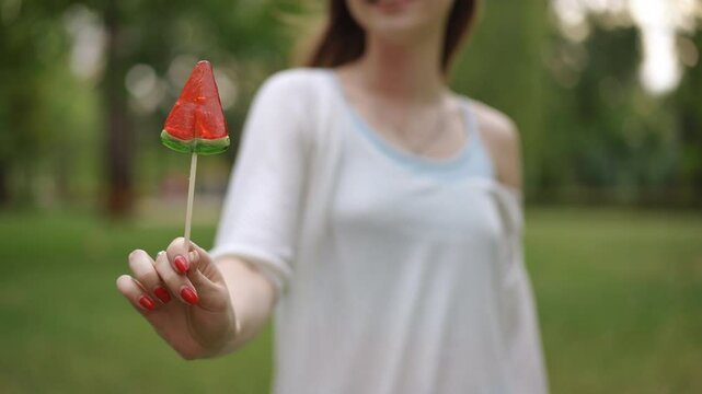 A young girl joyfully enjoys a delightful and colorful watermelon lollipop while outdoors in a lively green setting