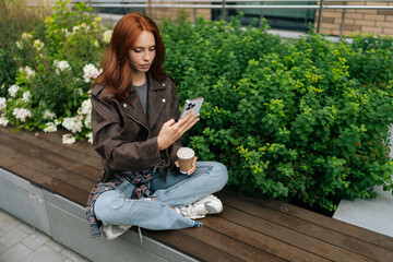 Charming young woman in leather jacket enjoying coffee break sitting on city bench, checking phone. Pretty redhead female using smartphone holding coffee outdoors.