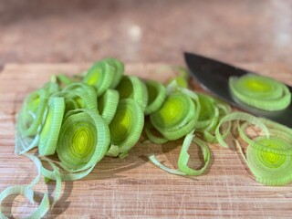 Detailed view of thinly sliced green leeks on a wooden cutting board, with the shiny chefs knife off to the side. The food preparation area is an example of a home kitchen setting. 