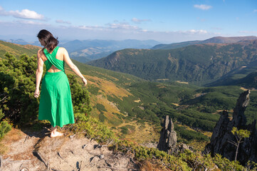 A woman in a flowing green dress stands barefoot on a rocky mountain peak, surrounded by stunning landscapes under a clear blue sky. Nature, freedom, and exploration