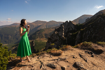 A woman in a flowing green dress stands barefoot on a rocky mountain peak, surrounded by stunning landscapes under a clear blue sky. Nature, freedom, and exploration