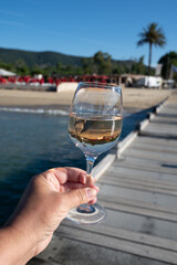 Hand with glass of cold rose wine from Provence and wooden yacht boota pier on white sandy beach Plage de Pampelonne near Saint-Tropez, summer vacation in France