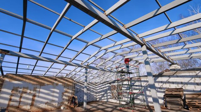 Footage at the spacious attic of the construction. Worker welds the metal balks of the future roof. Low angle view.