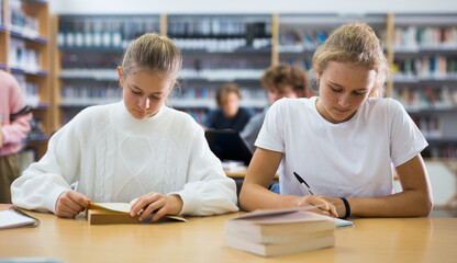Teenager girls sitting at table in reading room in library and doing homework together.