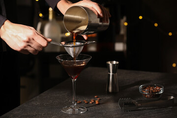 Bartender making delicious espresso martini at dark table against blurred lights, closeup