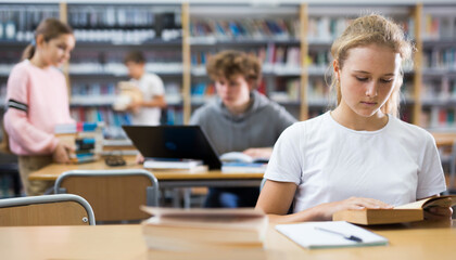 Portrait of a fifteen-year-old schoolgirl sitting at a desk in the school library, attentive reading a book
