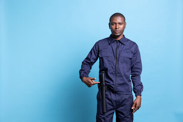 Fototapeta premium Male security guard pointing to his baton to ensure protection on duty, standing against blue background in patrolling uniform. Confident serious agent presenting his weapon, public safety.