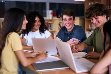 Group of young high school students doing classwork together using laptop sitting in library. Happy diverse Generation z classmates meeting brainstorming studying with computers and notebooks