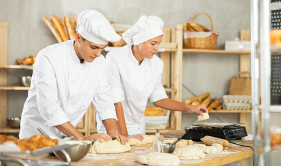 Positive male baker kneading dough and girl cutting it and weighing on scales in bakery