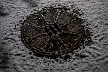 A close-up view of a round manhole cover covered by fresh snow. The snow contrasts with the wet, textured surface of the metal cover, creating a striking winter detail illuminated in dim light