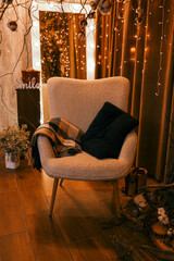 A cozy interior with a soft armchair, a blanket and pillows against the background of a warm garland of lighting. Elements of winter decor create an atmosphere of coziness and celebration