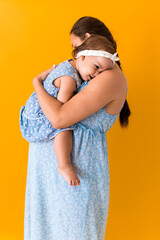 Motherhood, hot summer - portrait young pregnant happy smiling joy mother woman in blue dress holding little baby daughter toddler sibling bare feet in arms looking into eyes kiss on yellow background