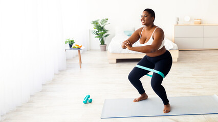 Fototapeta premium Chubby millennial black woman squatting with elastic band during domestic workout, free space. Overweight African American female training leg muscles, doing sports at home