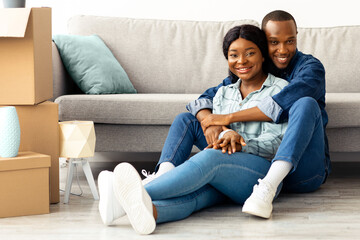 Happy african american couple sitting on floor in room with cardboard boxes on moving day, cheerful black spouses embracing and smiling to camera, celebrating relocating to new apartment, free space