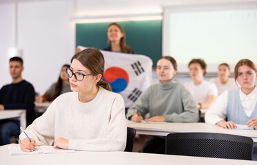 Students sit at desks, teacher stands behind with flag of South Korea