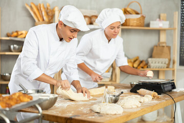 Young baker girl and guy work near work table in kitchen, during knead dough, cut pieces of dough from large lump and put them on scales. Employee weighs dough pieces, shape balls for baking buns