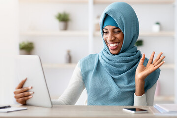 Online Communication. Cheerful Black Muslim lady In Hijab Making Video Call With Digital Tablet While Sitting At Desk In Home Office, African Islamic Freelancer Lady Waving Hand At Web Camera