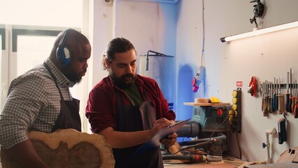 Craftsperson and colleague look over technical schematics to make commissioned wooden object. Manufacturer and coworker analyze blueprints to execute furniture assembling woodworking project, camera B