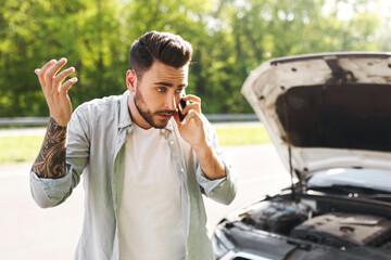 Upset young Caucasian man standing near his broken car with open hood, experiencing engine failure, having mobile phone conversation, calling breakdown road service on cellphone