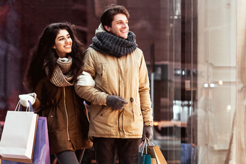 Christmas Sale. Couple Doing Winter Shopping Carrying Shopper Bags Walking Along Stores In Night City. Free Space, Panorama